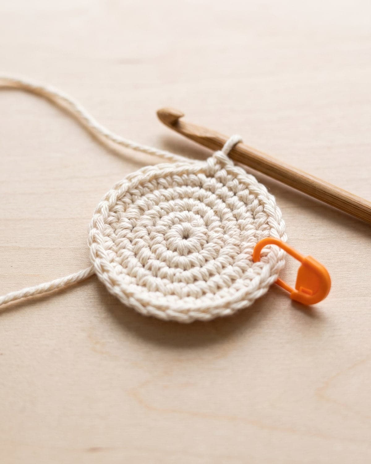 Small flat circle of cream crochet with an orange stitch marker and a wooden hook