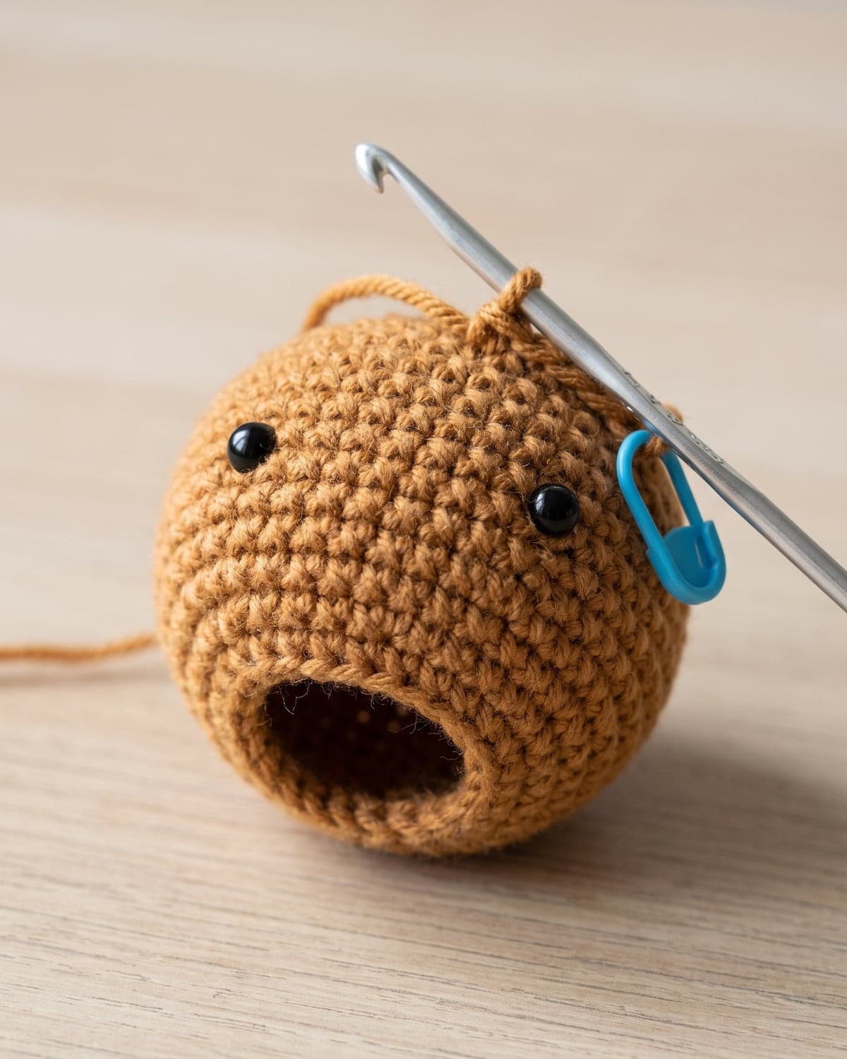 Unfinished caramel crochet bunny head with safety eyes, an opening, a hook and a blue stitch marker