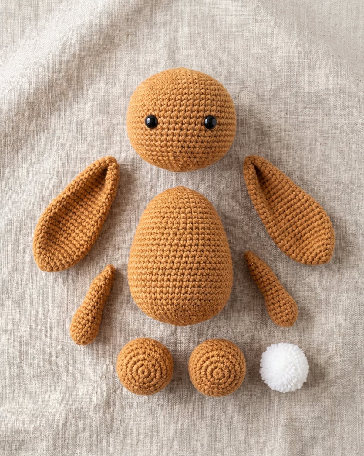 Crochet bunny pieces laid out on linen: head, body, two long ears, two arms, two feet and a white pompom tail