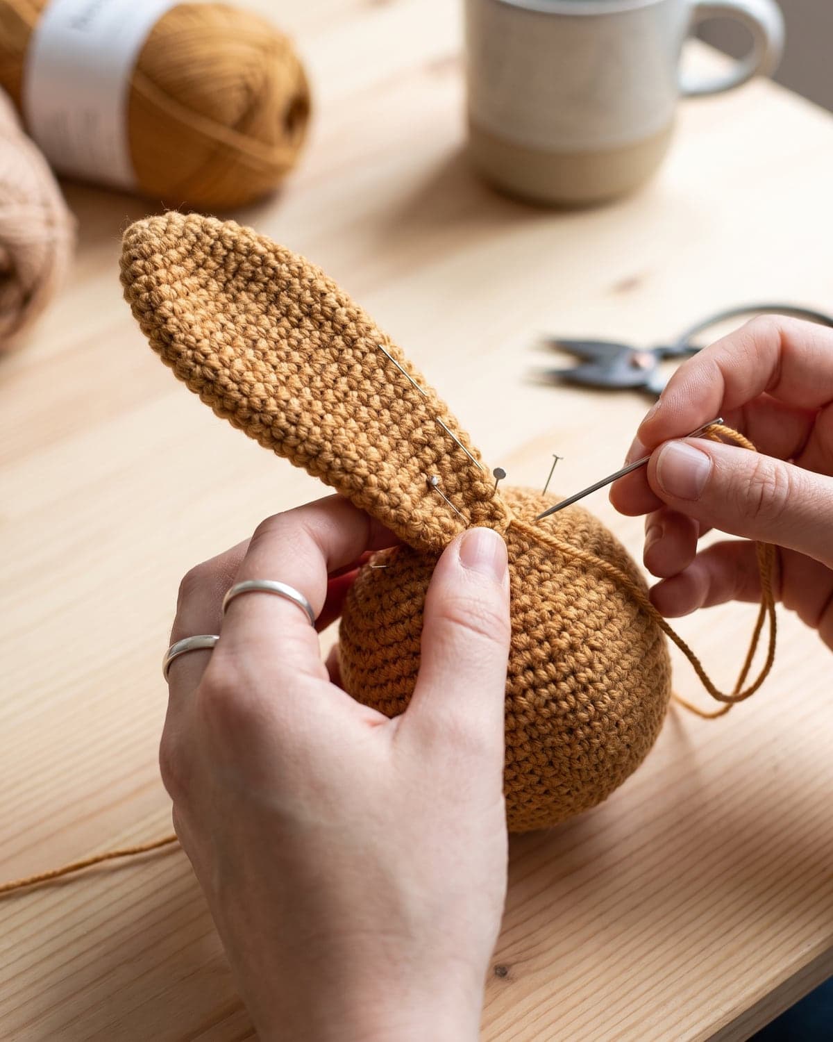 Hands sewing a long ear onto the head of a caramel crochet bunny with pins and a needle