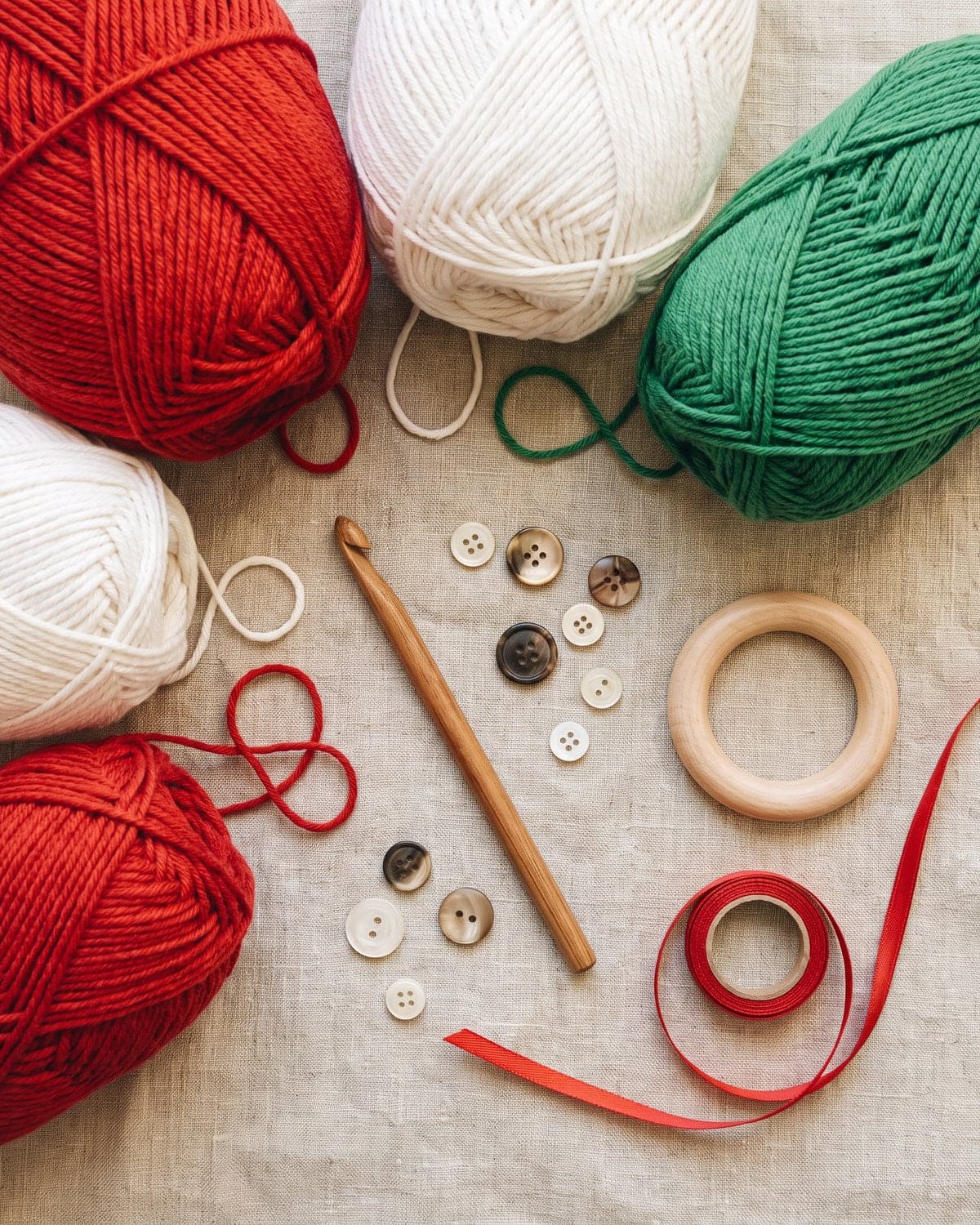 Red, white and green cotton yarn with a wooden crochet hook, buttons, a wooden ring and red ribbon on linen