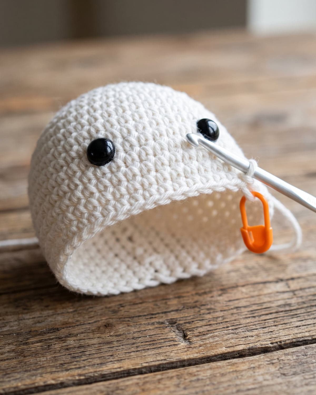 Unfinished white crochet ghost body with black safety eyes, a hook in the loop and an orange stitch marker