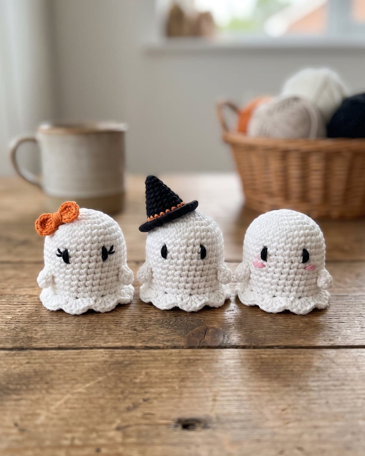 Three white crochet ghosts on a wooden table, one with an orange bow, one in a witch hat, one with pink cheeks