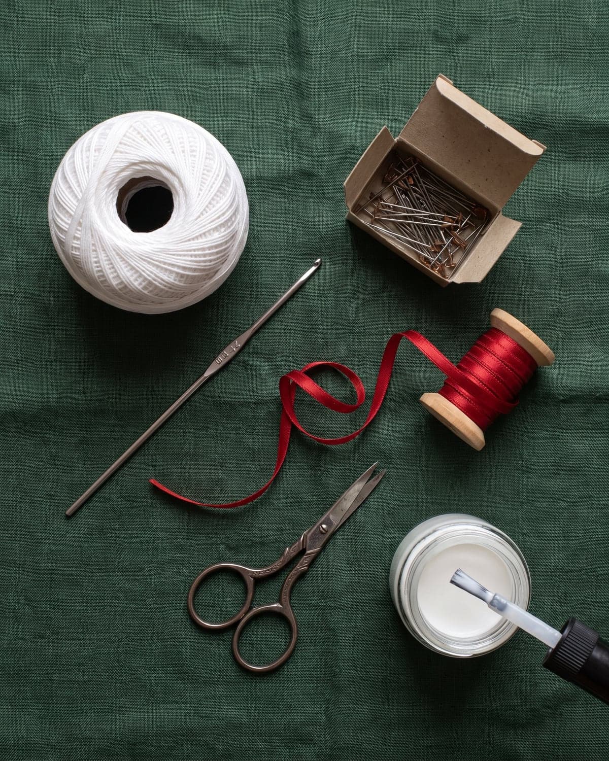 White cotton crochet thread, a steel hook, pins, red ribbon, scissors and a jar of glue with a brush on green cloth