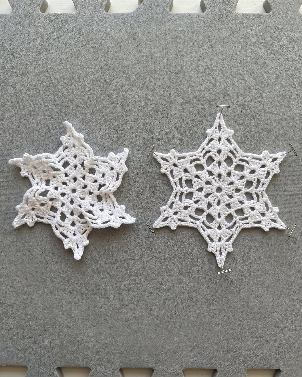 A curled white crochet snowflake beside one pinned out flat on a grey blocking mat