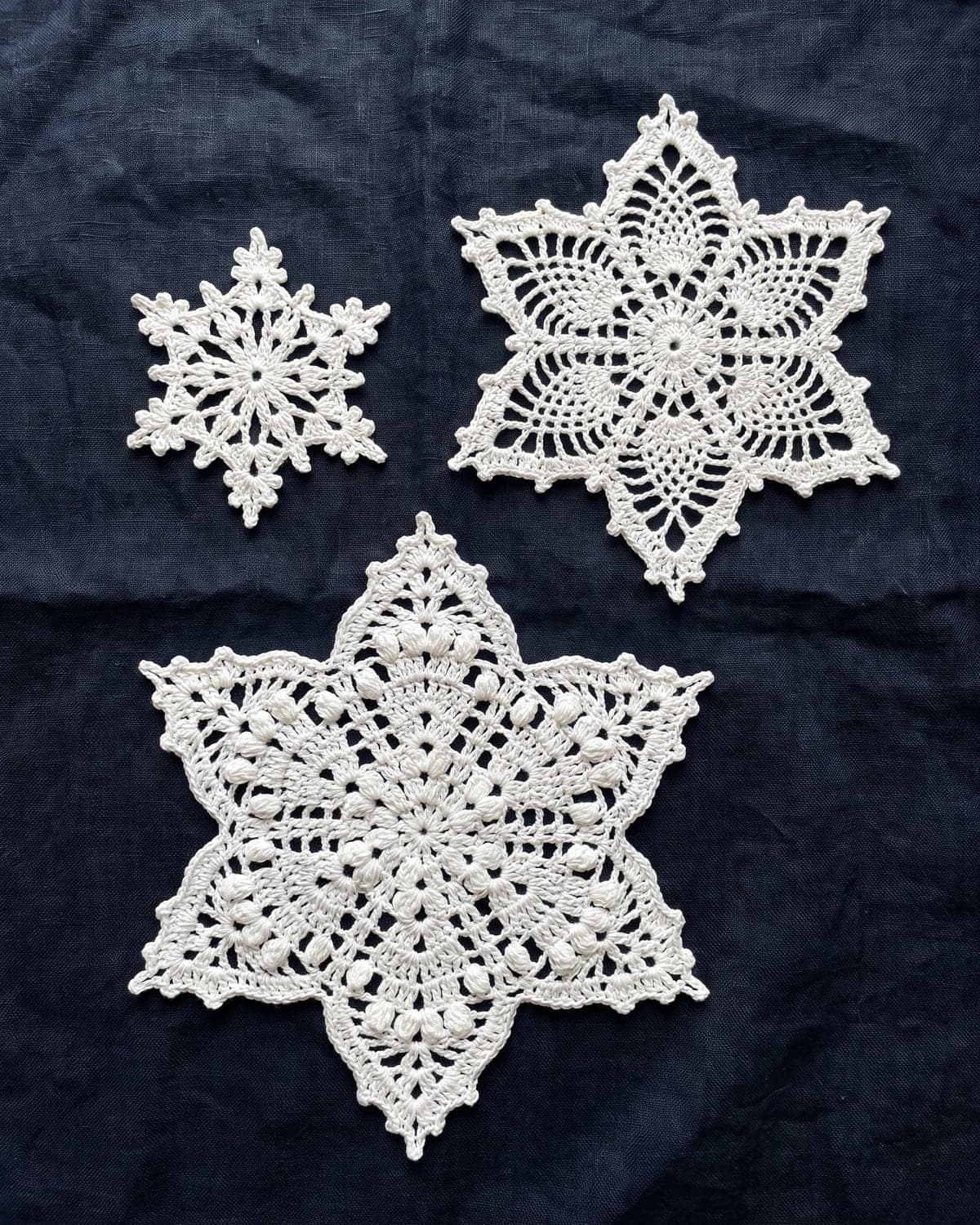 Three white crochet lace snowflakes in different sizes on dark navy linen