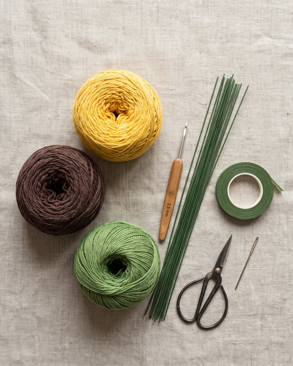 Yellow, brown and green cotton yarn with a crochet hook, floral wire, floral tape, scissors and a needle on linen