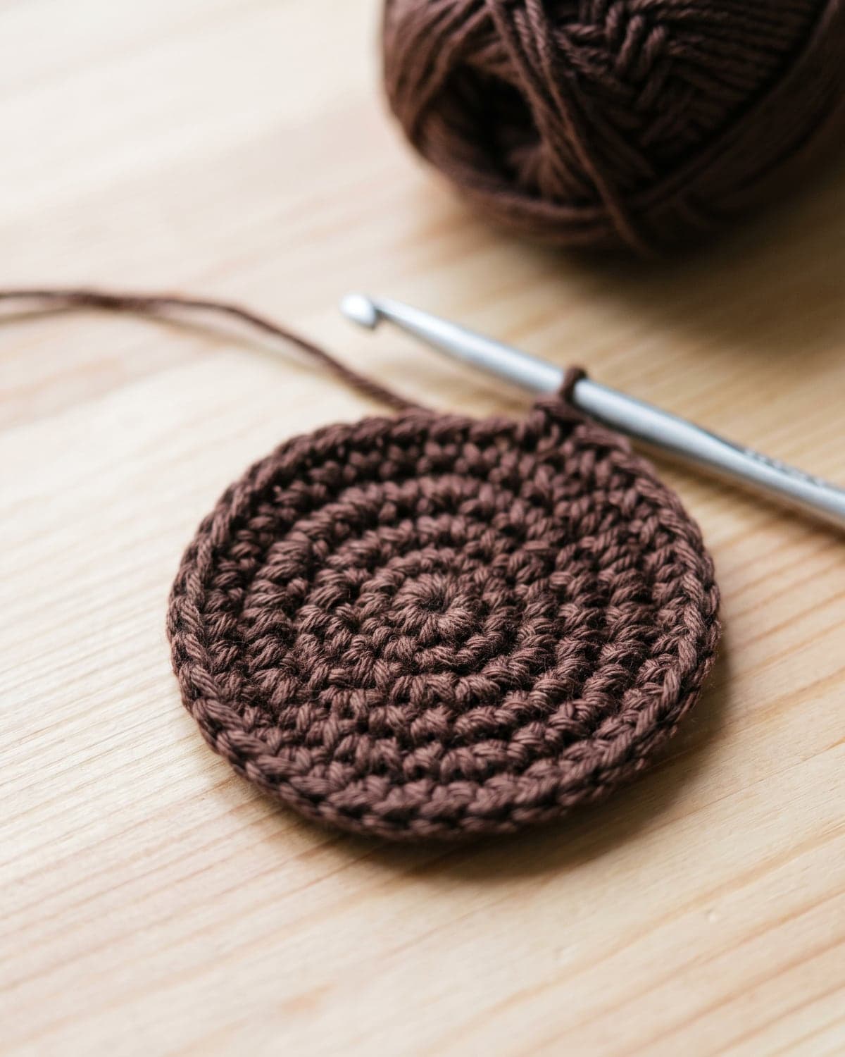 Flat round crochet disc in dark brown yarn with a hook in the loop, the centre of a sunflower