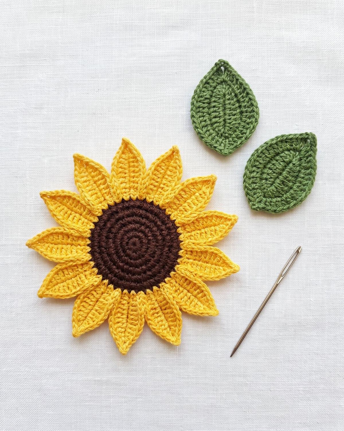 Finished flat crochet sunflower beside two green crochet leaves and a tapestry needle