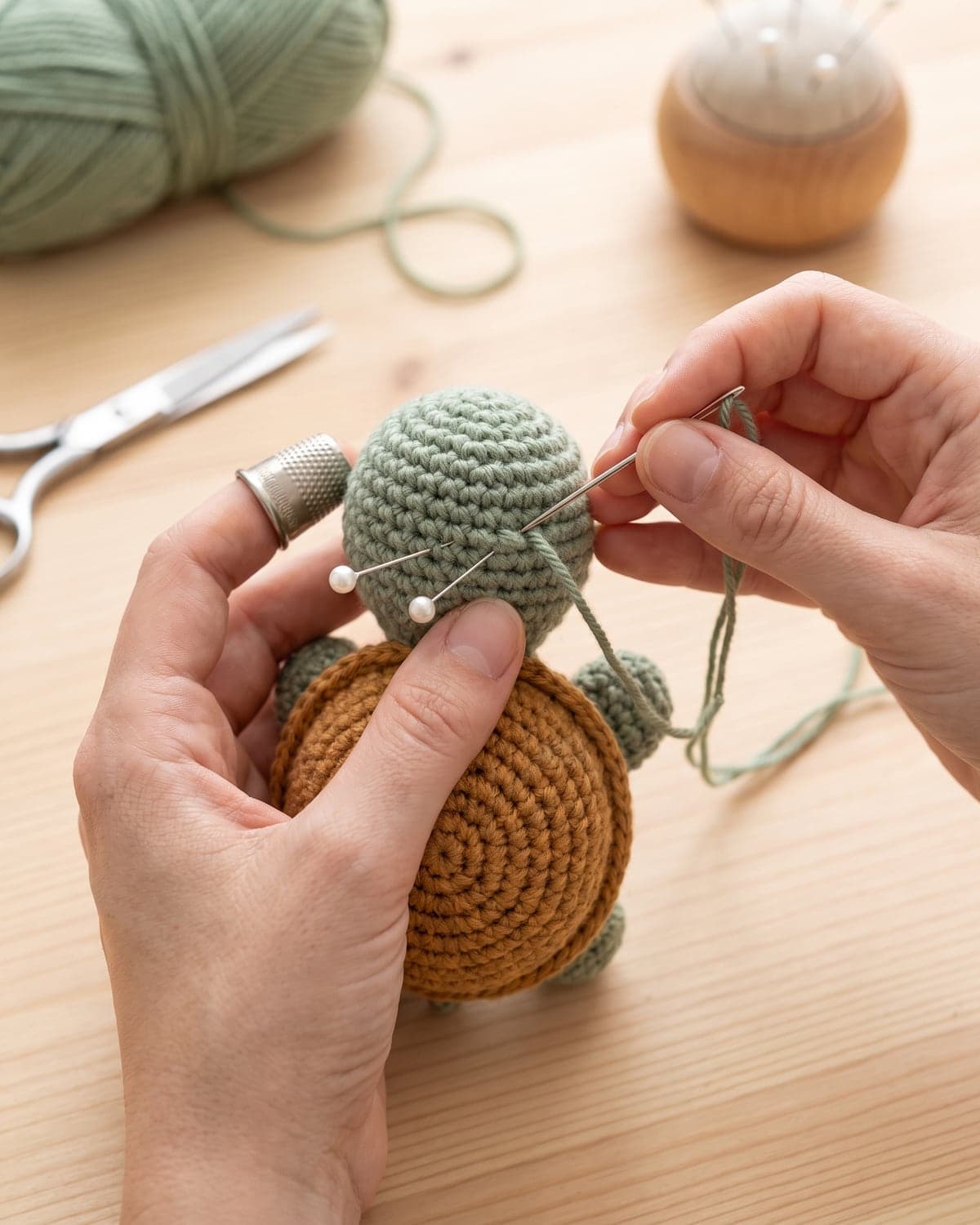Hands sewing the green head onto a crochet turtle, held in place with pins