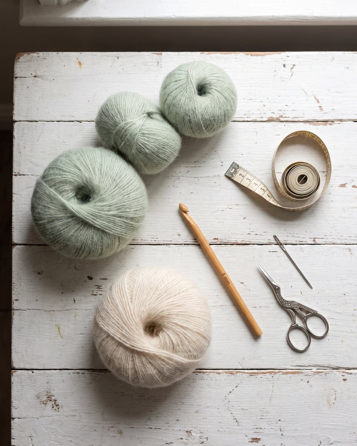 Balls of soft sage green and cream yarn with a crochet hook, tape measure, scissors and needle on white wood