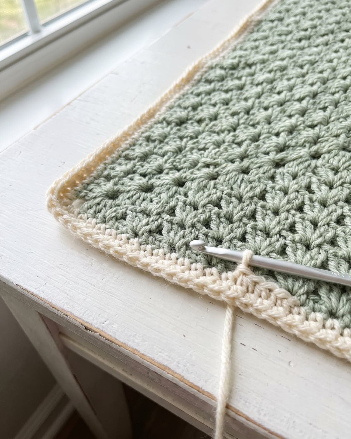 Cream crochet border being worked around the corner of a sage green baby blanket