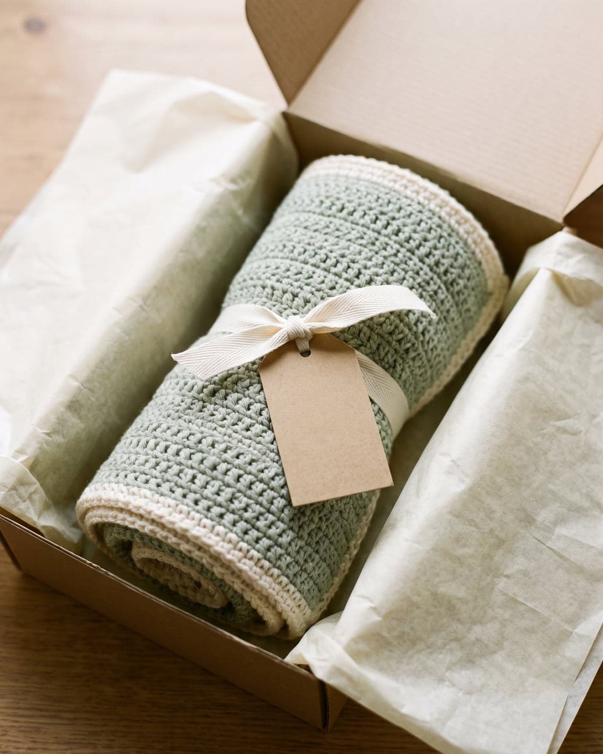 Sage green crochet baby blanket rolled and tied with cream ribbon and a blank tag in a gift box