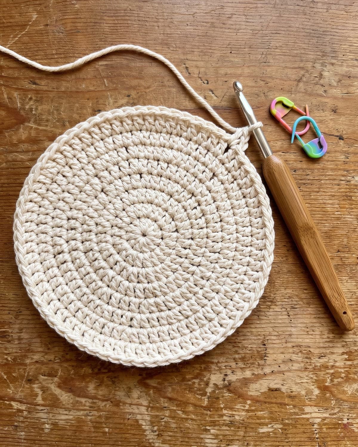 Flat round crochet basket base in cream cotton with a crochet hook and stitch markers on a wooden table