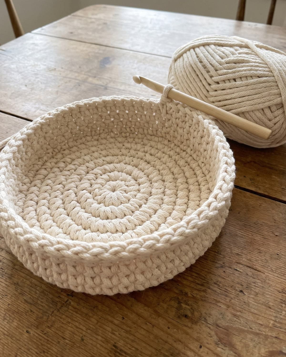 Cream crochet basket in progress with low walls rising from the round base and the hook still attached