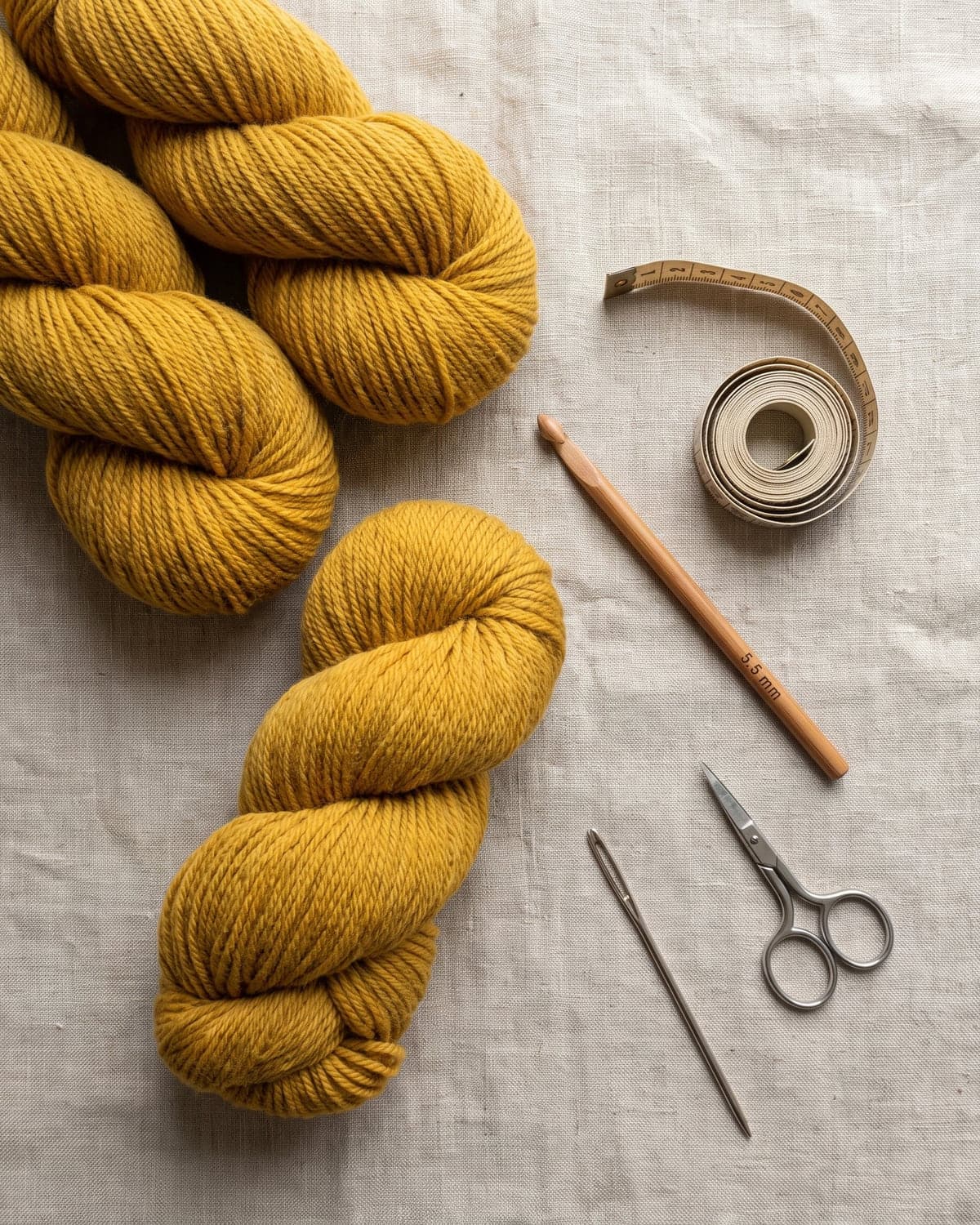 Skeins of mustard yellow yarn with a wooden crochet hook, tape measure, scissors and a needle on linen