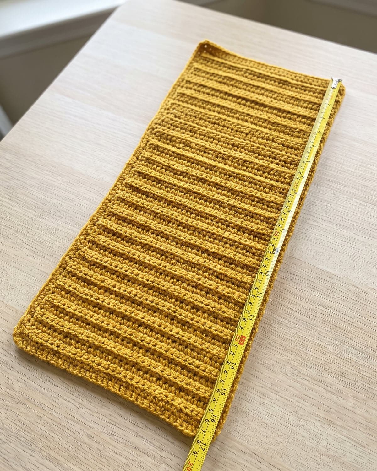 Flat ribbed mustard crochet rectangle on a table with a yellow tape measure along its edge