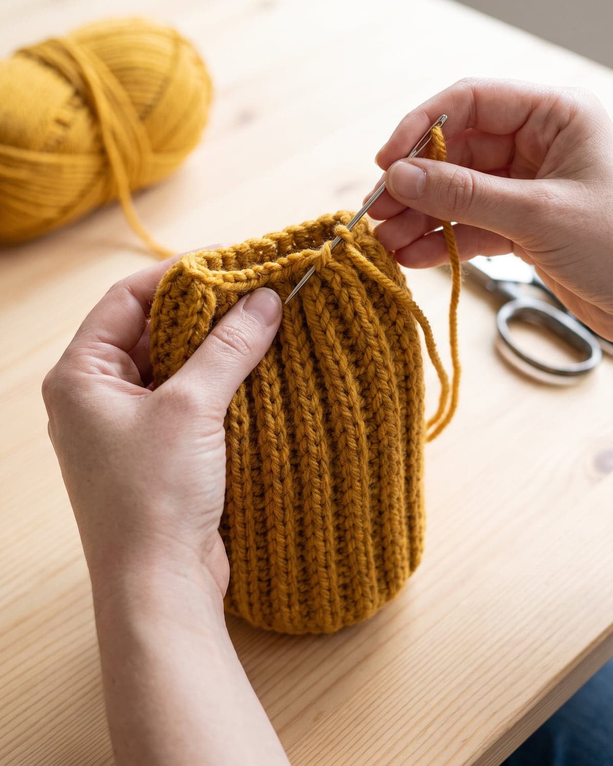 Hands gathering the top of a mustard ribbed crochet tube with a needle to close a beanie