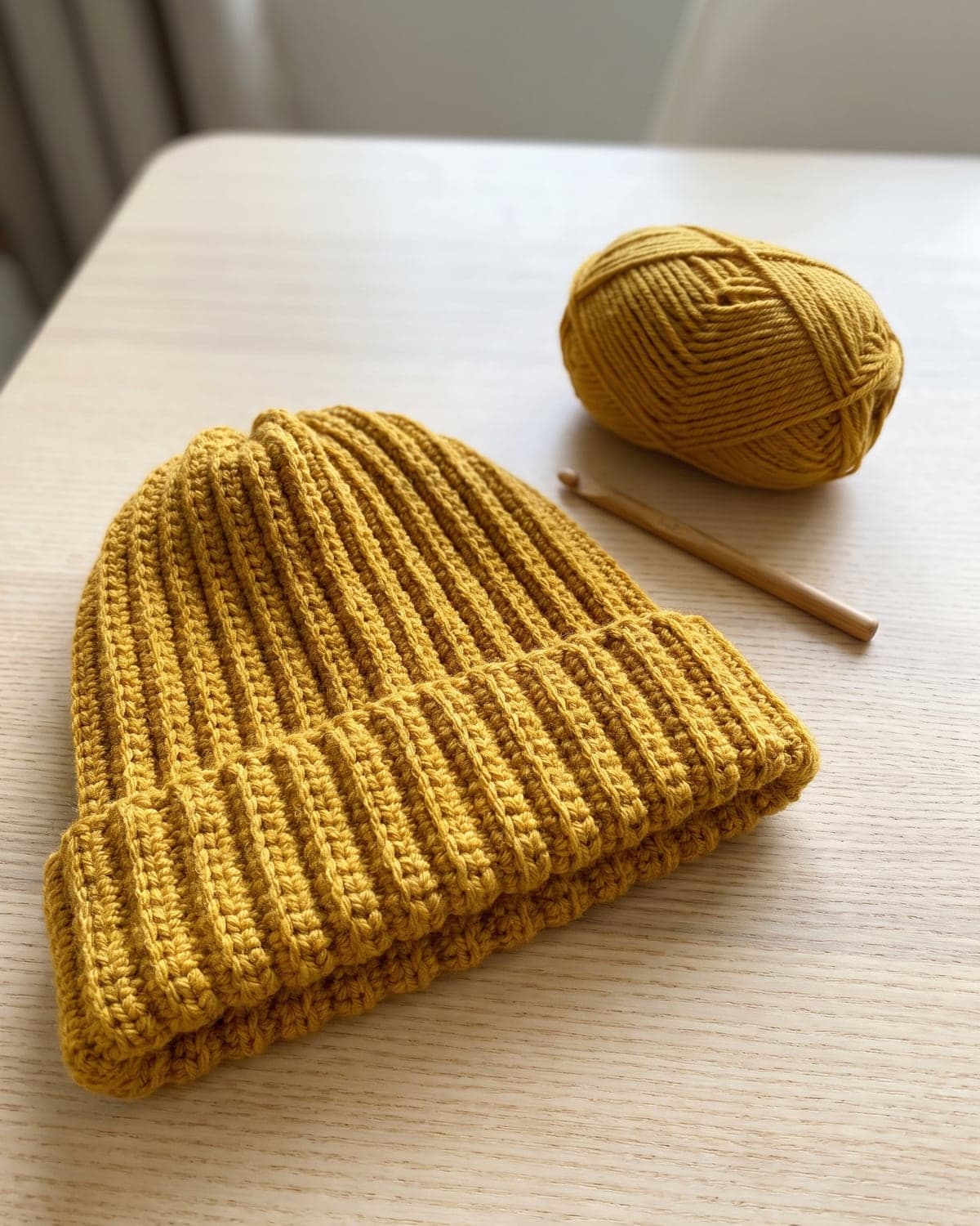 Mustard yellow ribbed crochet beanie with a folded brim beside a ball of yarn and a wooden hook