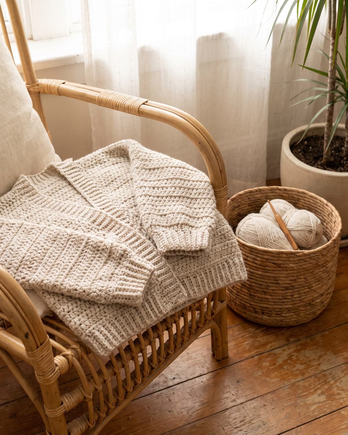 Folded oatmeal crochet cardigan on a rattan chair beside a basket of yarn