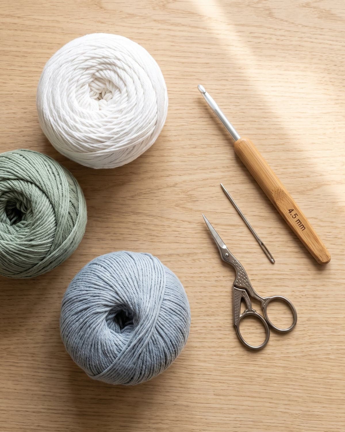 White, sage green and blue-grey cotton yarn with a crochet hook, scissors and a needle on a wooden table