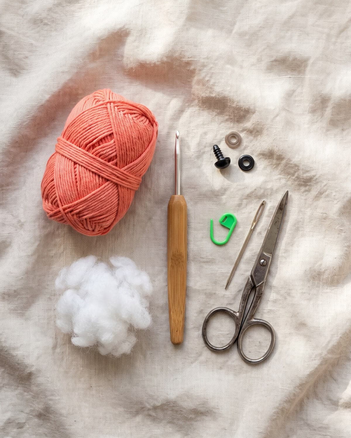 Coral cotton yarn, a crochet hook, a safety eye with washers, stuffing, a stitch marker, a needle and scissors on linen