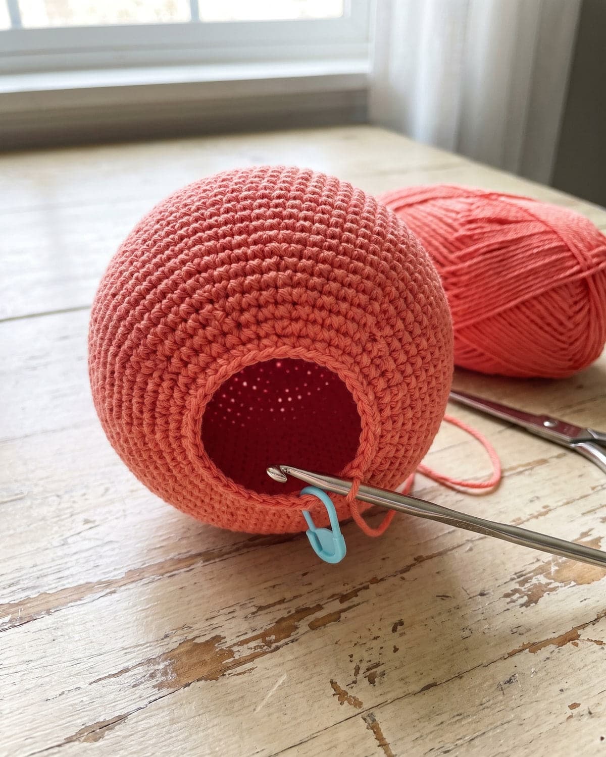 Unfinished coral crochet ball head with an opening, a crochet hook in the loop and a blue stitch marker