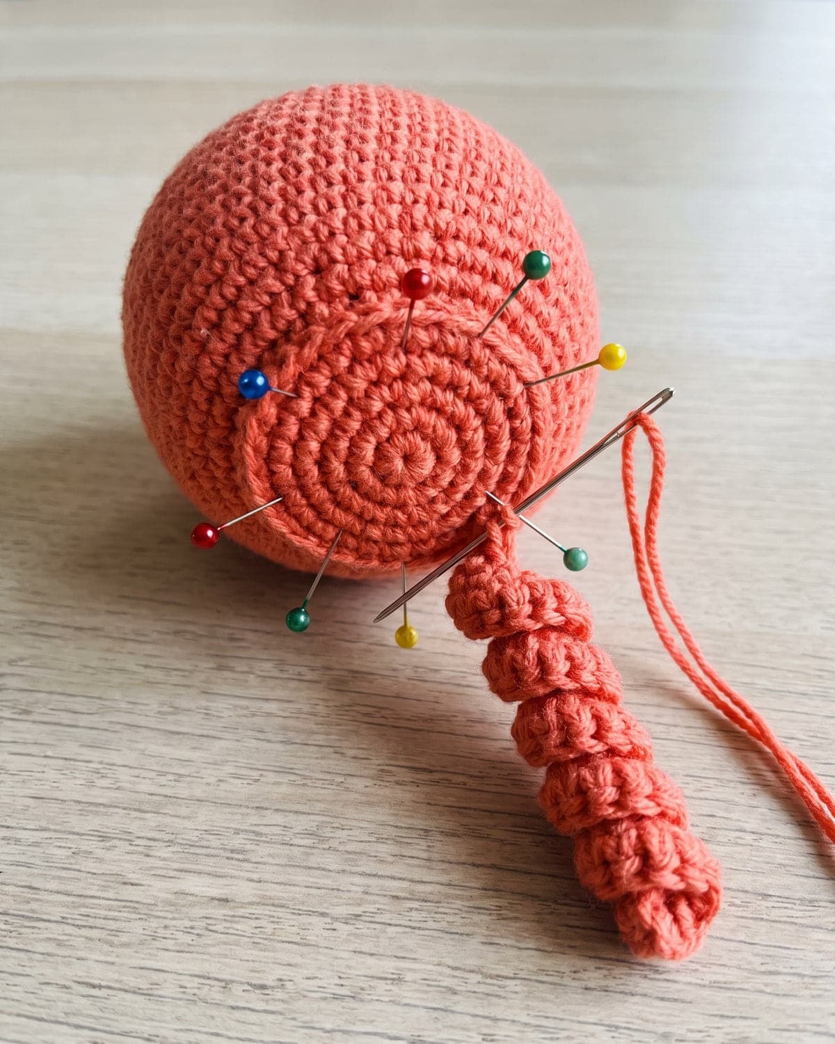 Coral crochet octopus head upside down with pins marking tentacle positions and one curly tentacle being sewn on