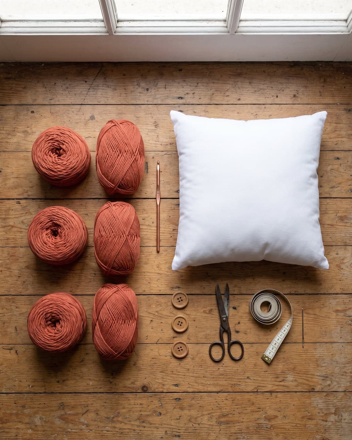 Terracotta yarn, a crochet hook, a white pillow insert, wooden buttons, scissors and a tape measure on a wooden floor