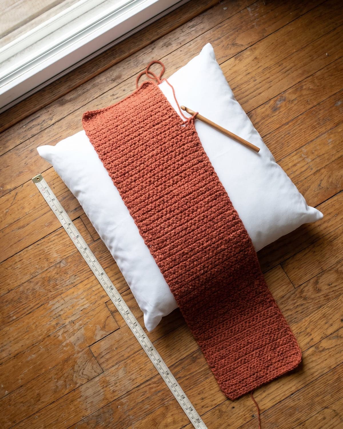 Long terracotta crochet rectangle laid over a white pillow insert with a tape measure beside it