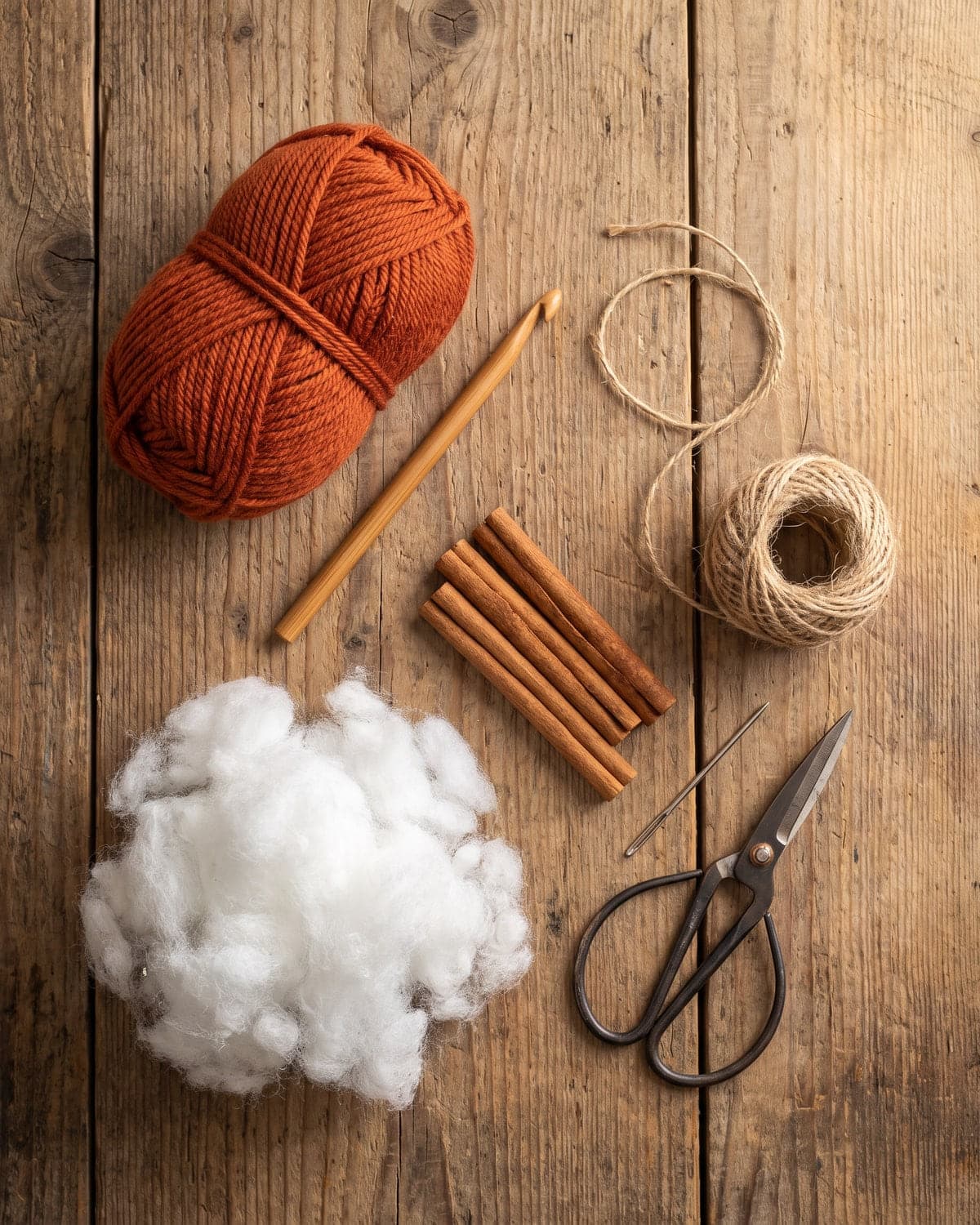 Burnt orange yarn, a crochet hook, stuffing, cinnamon sticks, jute twine and scissors on a wooden table