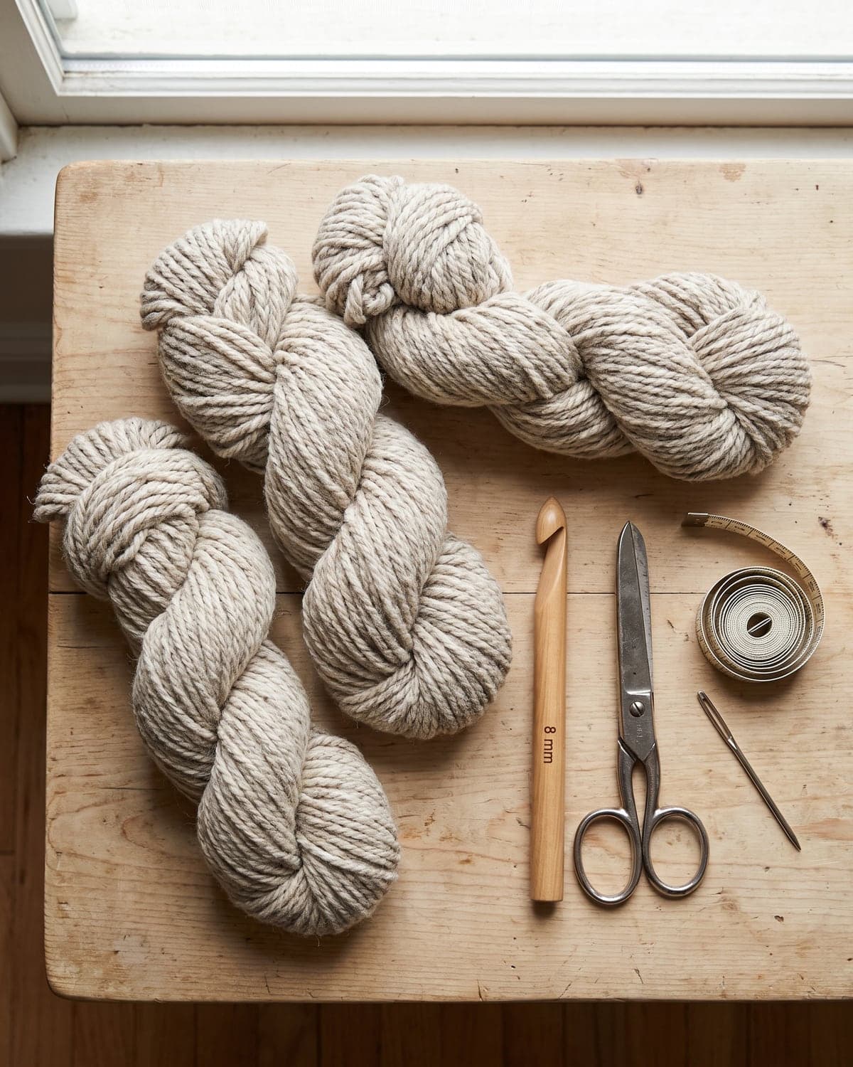 Three skeins of chunky oatmeal yarn with a large wooden crochet hook, scissors, tape measure and needle