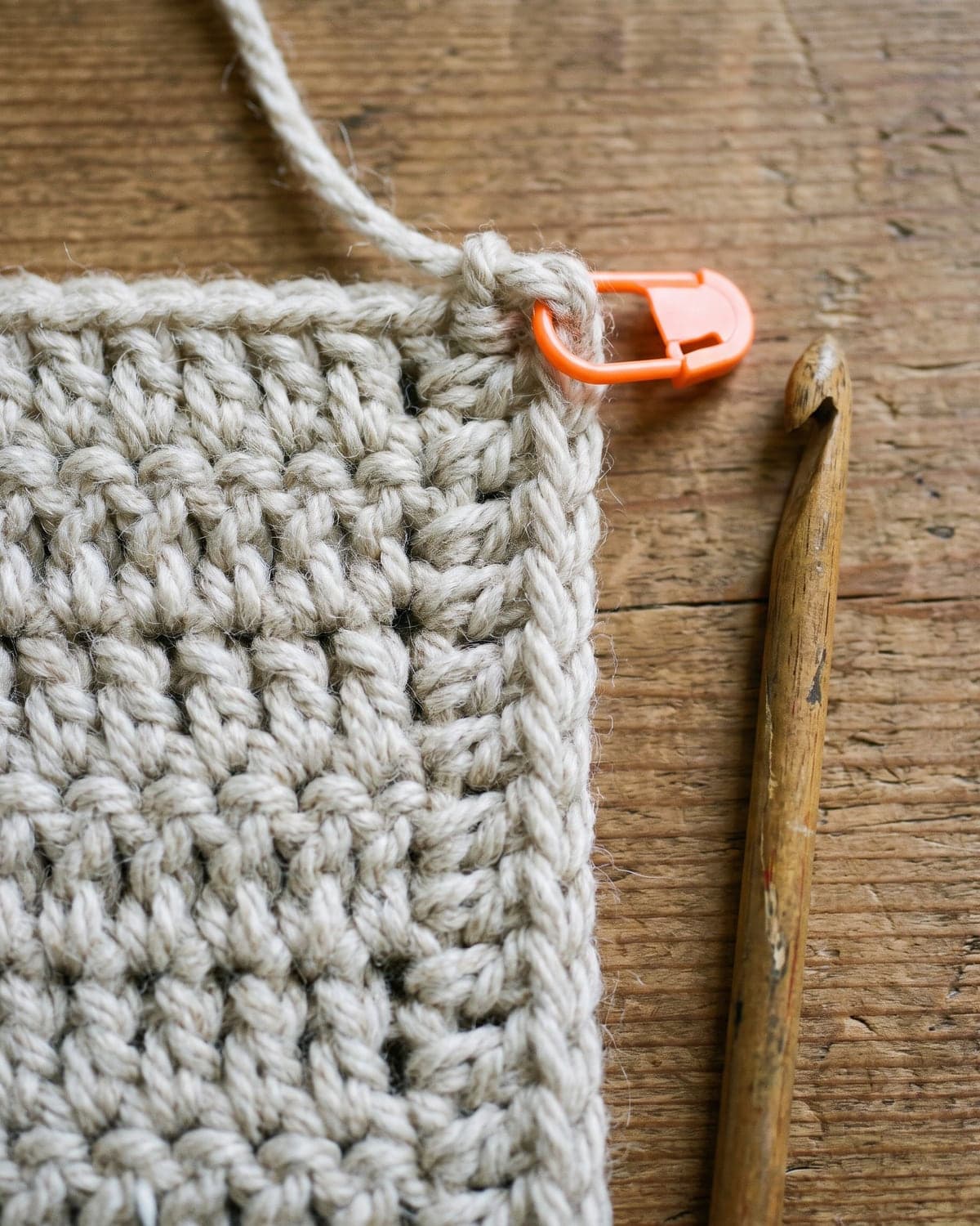 Straight edge of a chunky oatmeal crochet scarf with an orange stitch marker in the last stitch