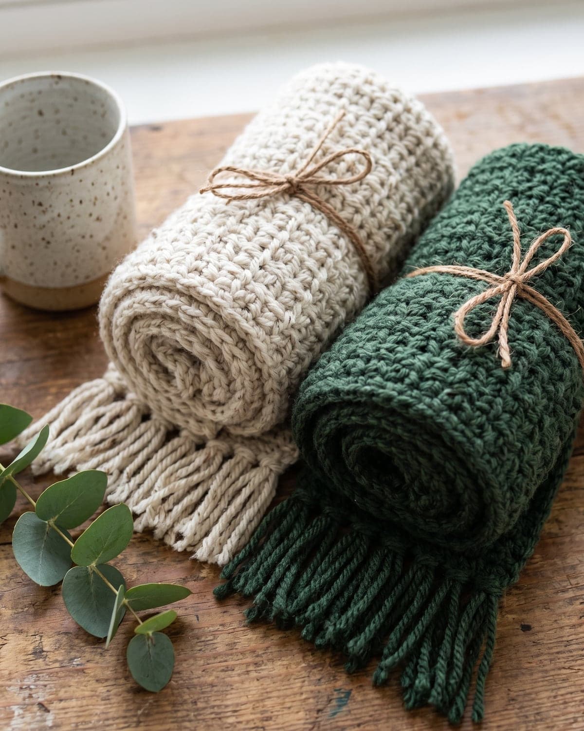Oatmeal and forest green crochet scarves with fringe, rolled and tied with twine on a wooden table