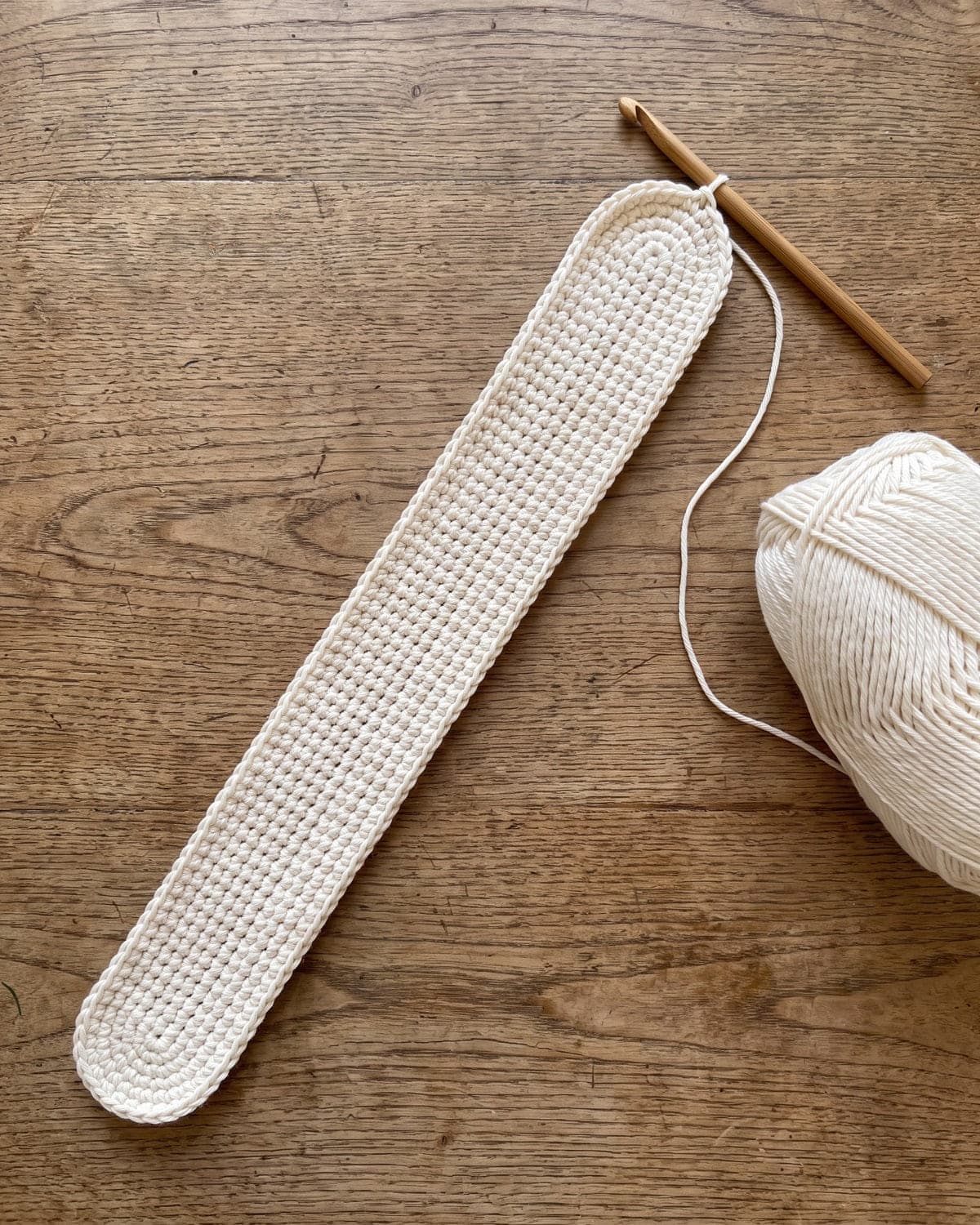 Long narrow crochet strip with rounded ends in cream cotton, the start of a bag base, with a hook and a ball of yarn