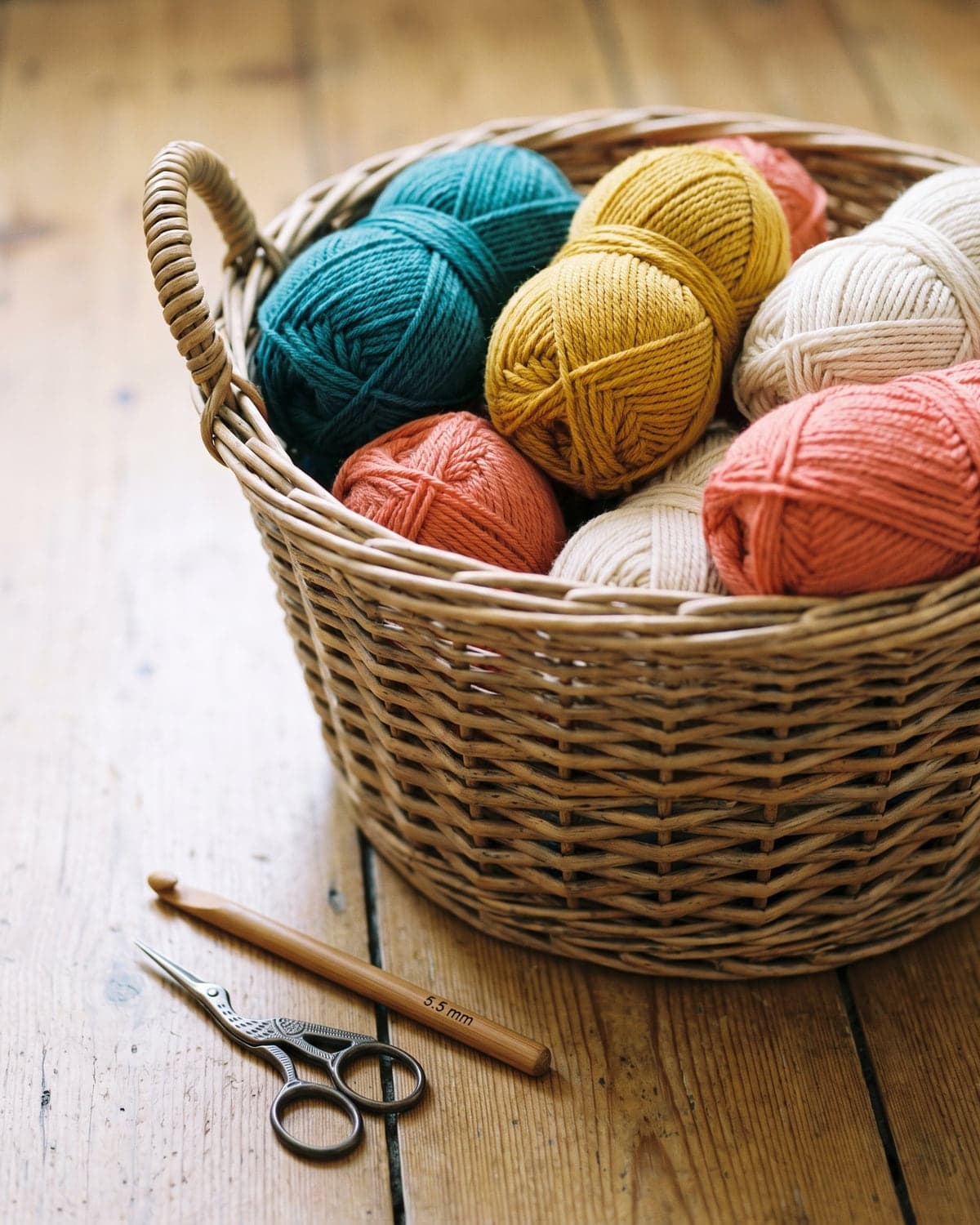 Basket of teal, mustard, cream and coral yarn with a crochet hook and scissors on a wooden floor