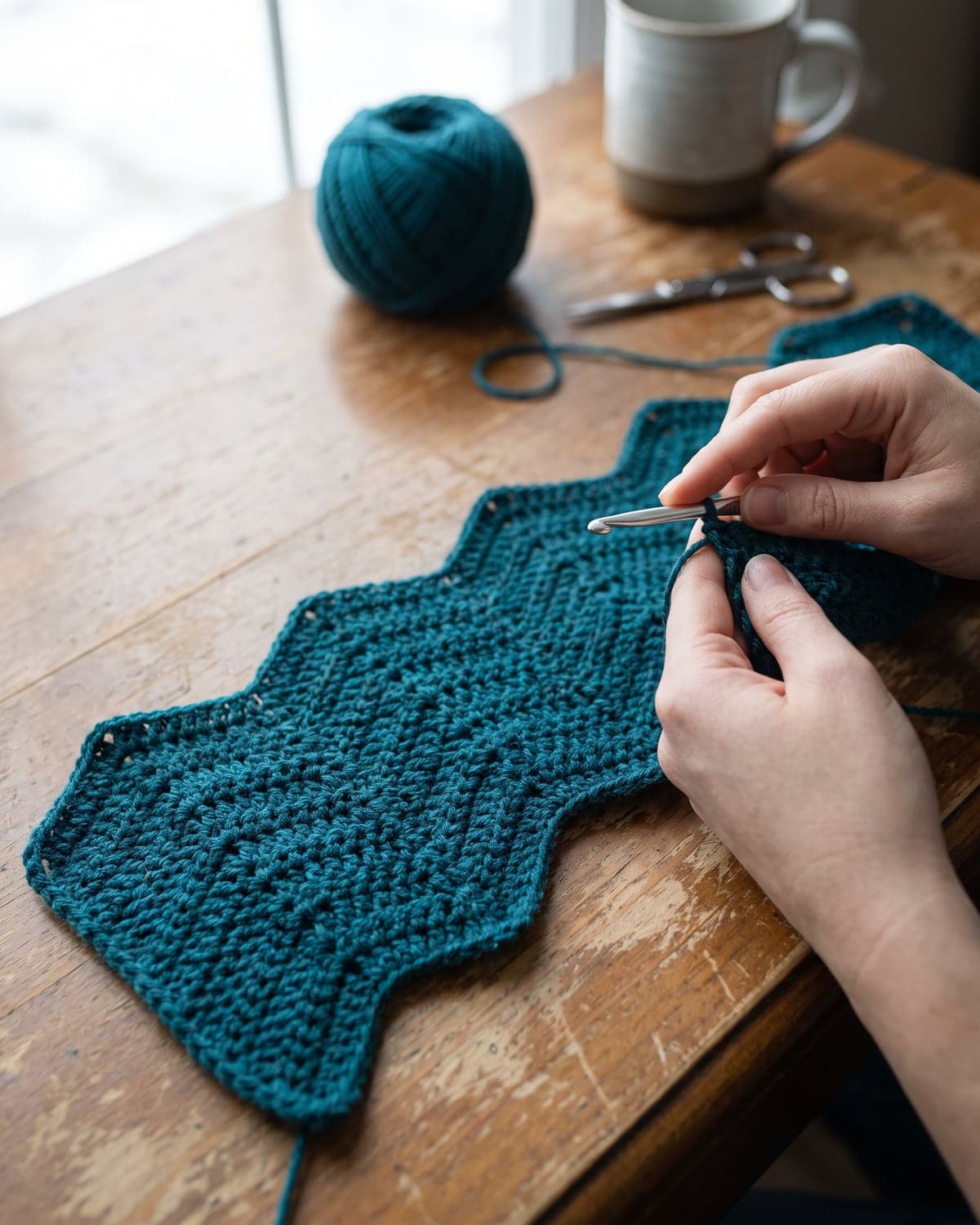 Hands crocheting the first zigzag rows of a teal ripple blanket on a wooden table