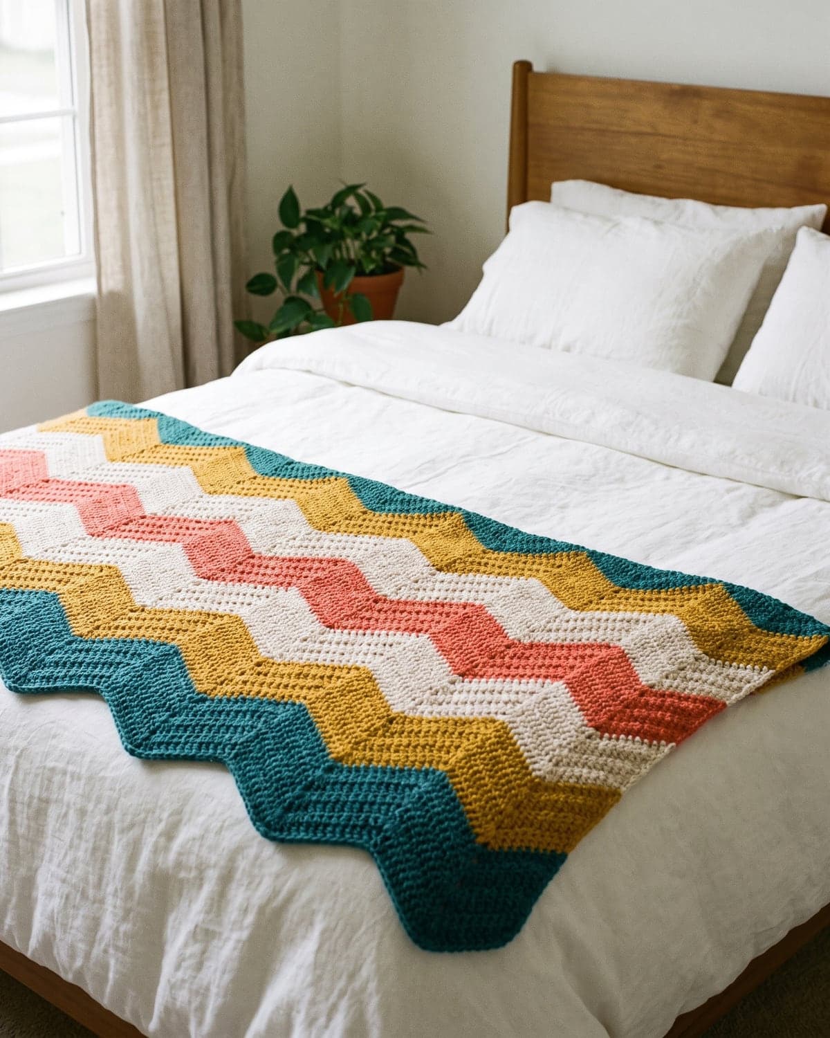 Striped crochet ripple blanket in teal, mustard, cream and coral laid across the foot of a white bed