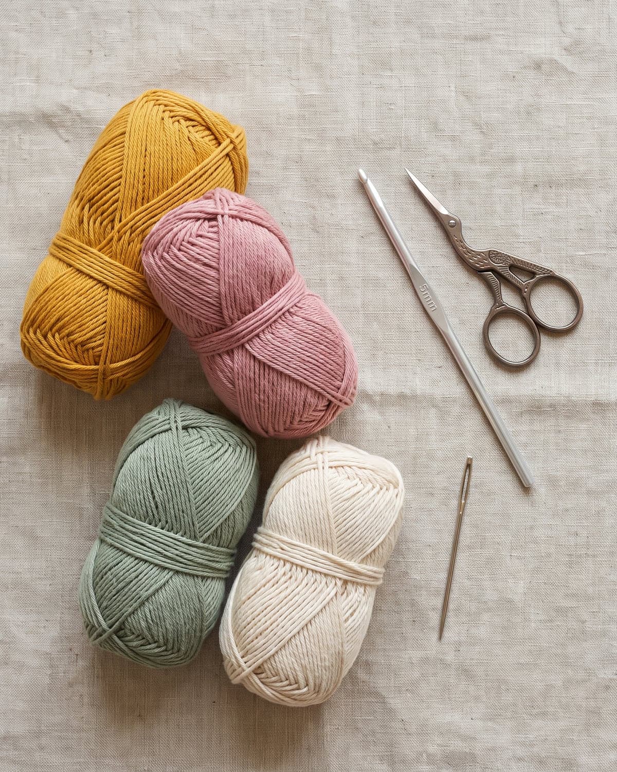 Mustard, pink, sage and cream cotton yarn with a crochet hook, scissors and a tapestry needle on linen
