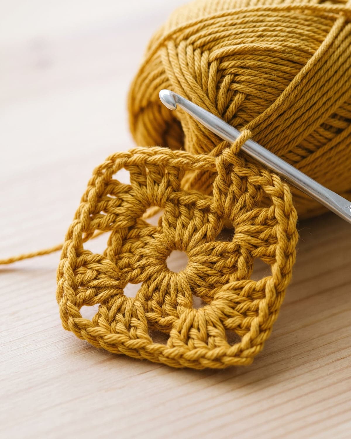 First round of a mustard crochet granny square with four clusters around a centre ring and a hook in the loop