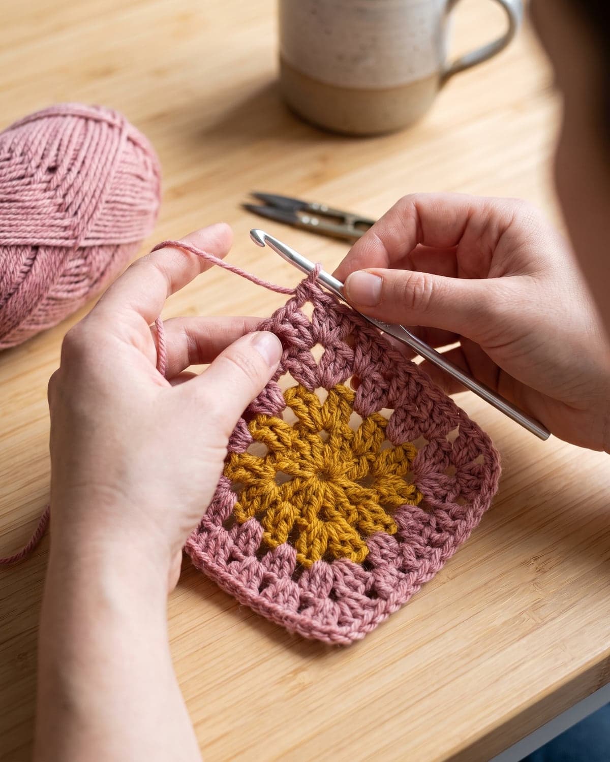 Hands crocheting the second round of a granny square in dusty pink around a mustard centre