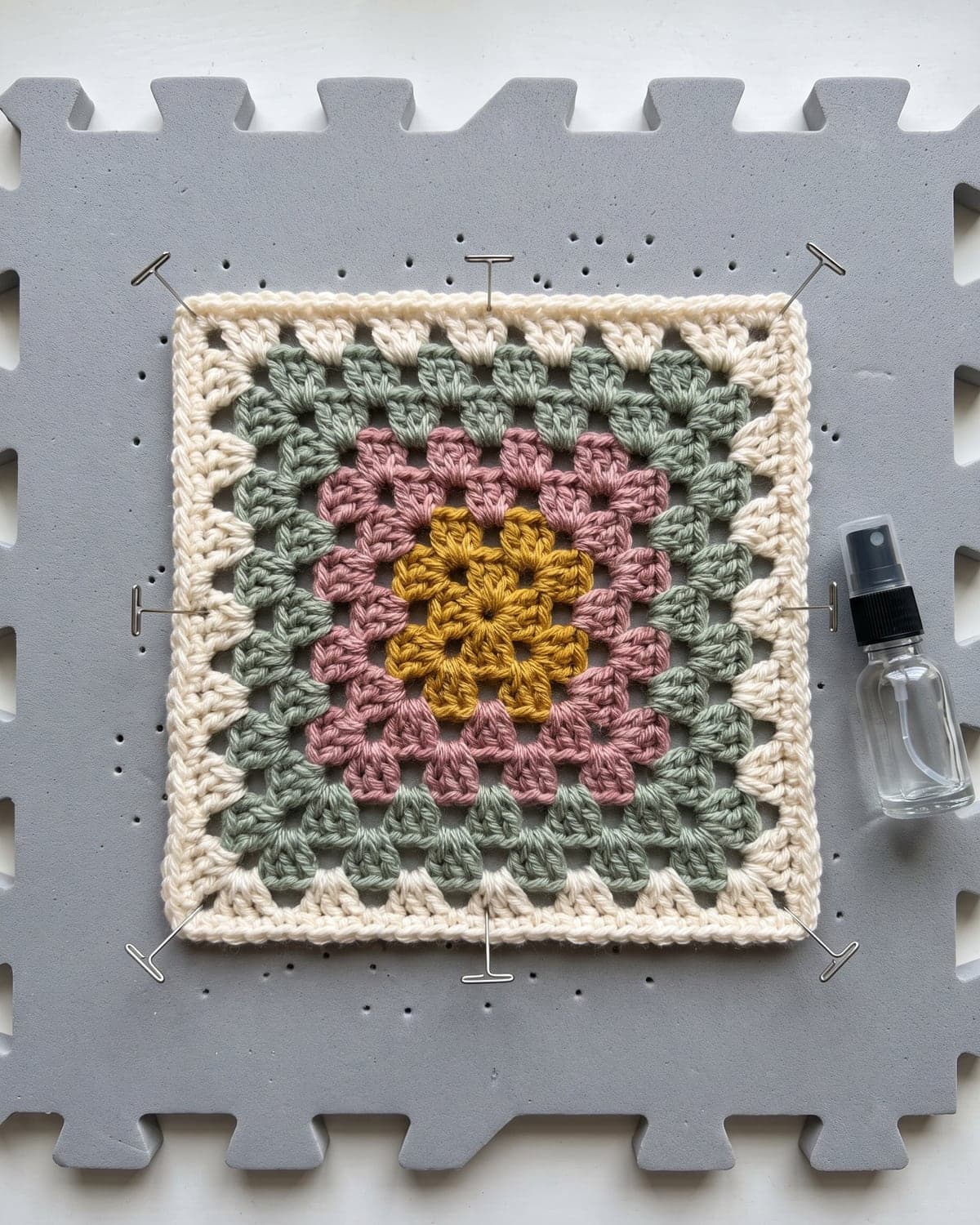 Four-round crochet granny square pinned flat on a grey blocking mat beside a spray bottle