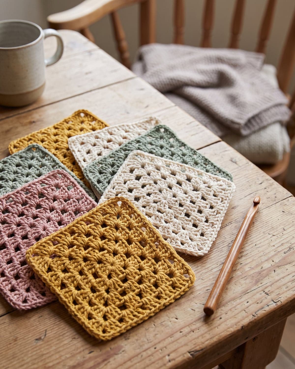 Classic crochet granny squares in mustard, dusty pink, sage green and cream overlapping on a wooden table with a crochet hook