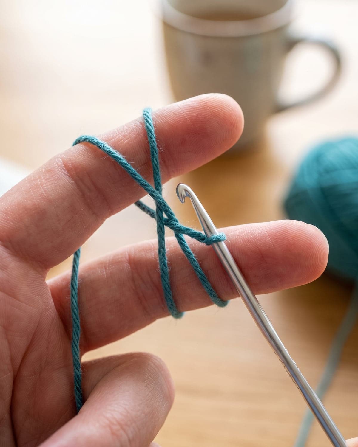 Teal yarn crossed over two fingers to start a crochet magic ring, with a hook sliding under the strand