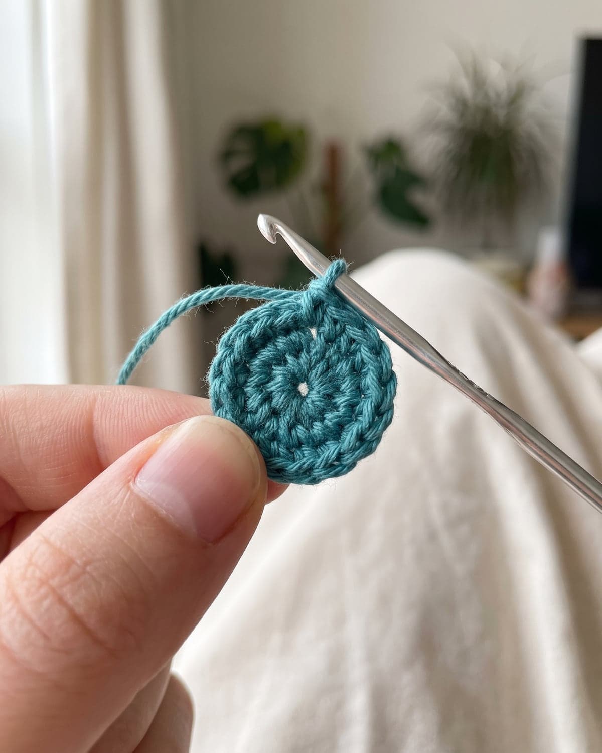 Small tightly closed teal crochet circle held between finger and thumb with a hook in the loop