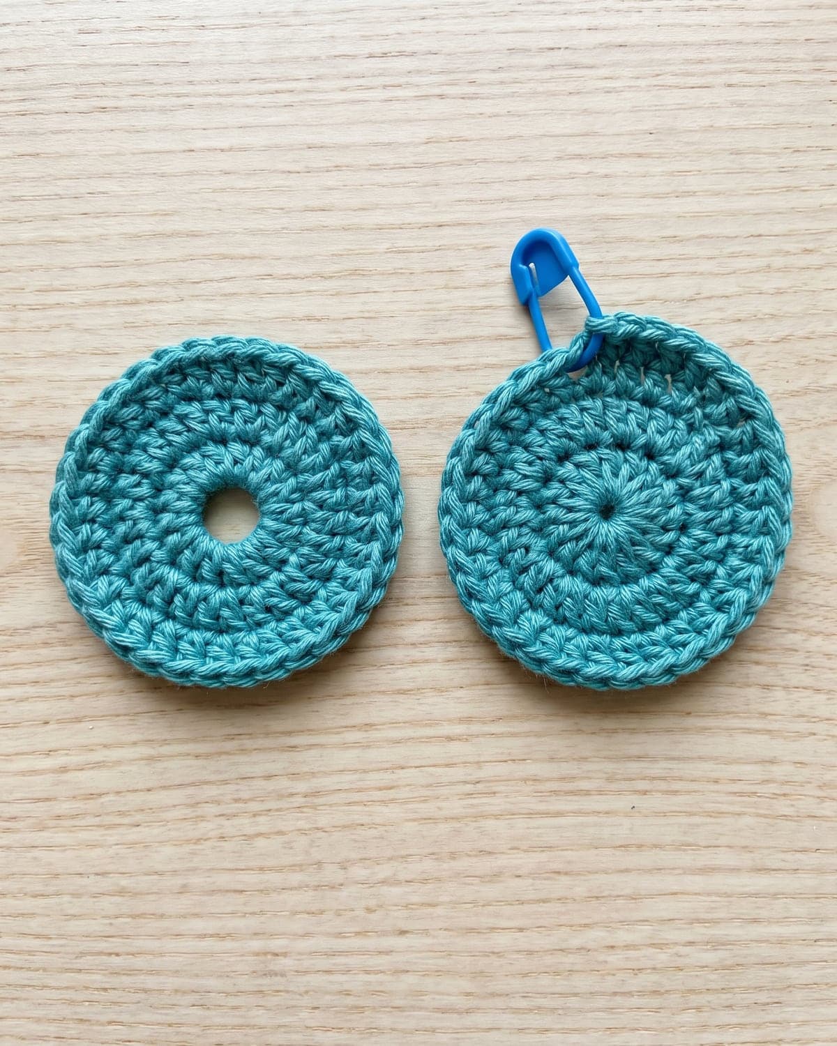 Two teal crochet circles side by side, one with a hole in the centre and one closed, with a blue stitch marker