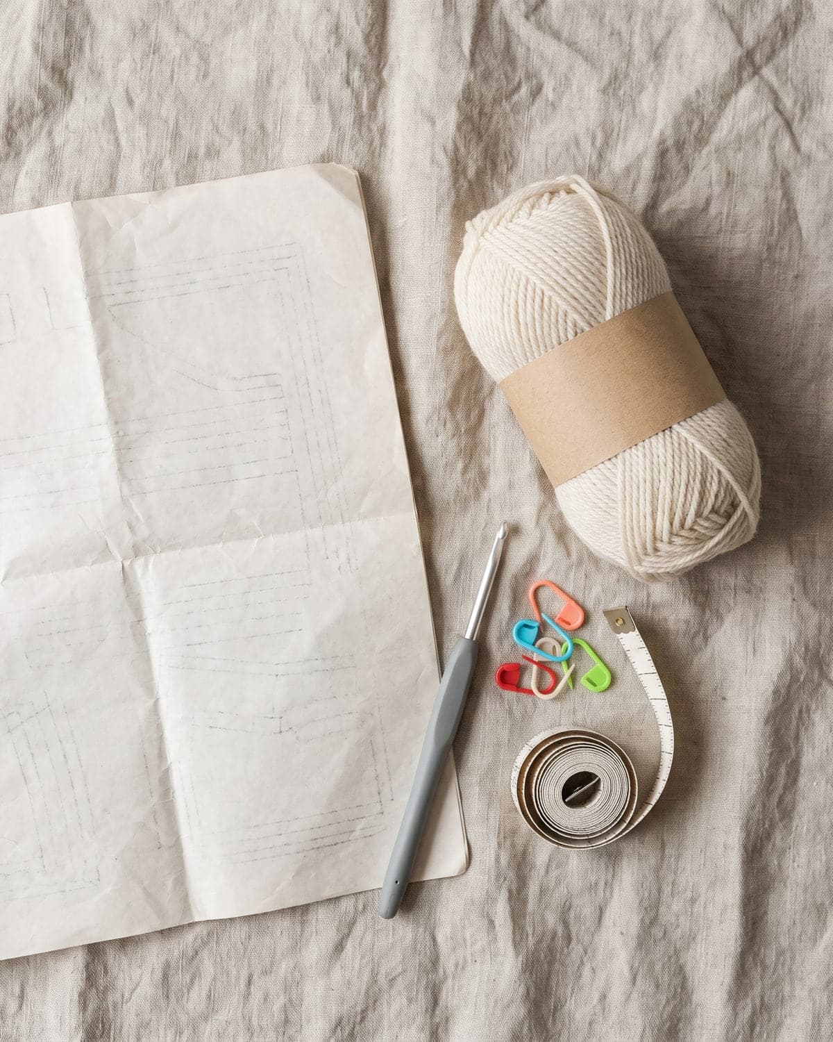 Folded pattern sheet, cream yarn with a blank label, a crochet hook, stitch markers and a tape measure on linen