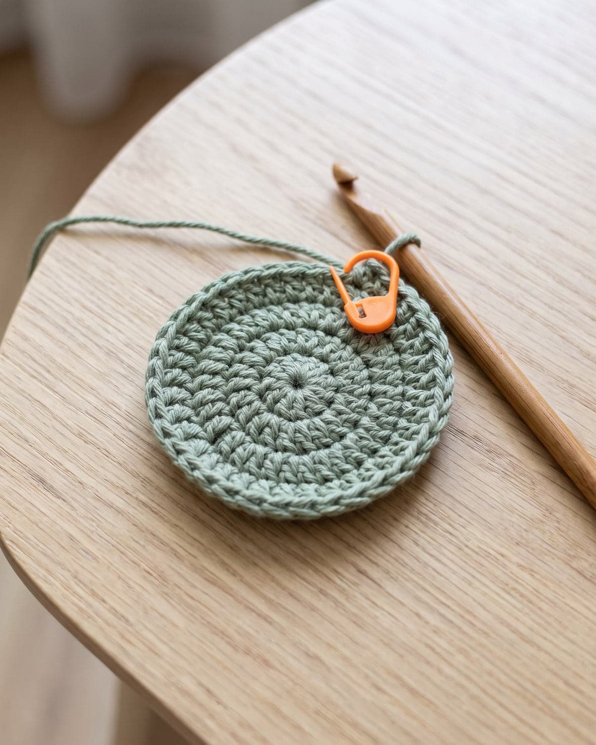Small flat crochet circle in sage green with an orange stitch marker and a wooden hook