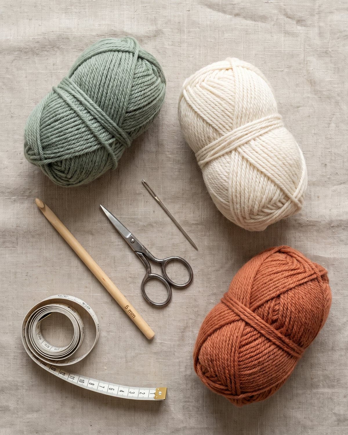 Sage, cream and terracotta yarn with a wooden crochet hook, scissors, a needle and a tape measure on linen