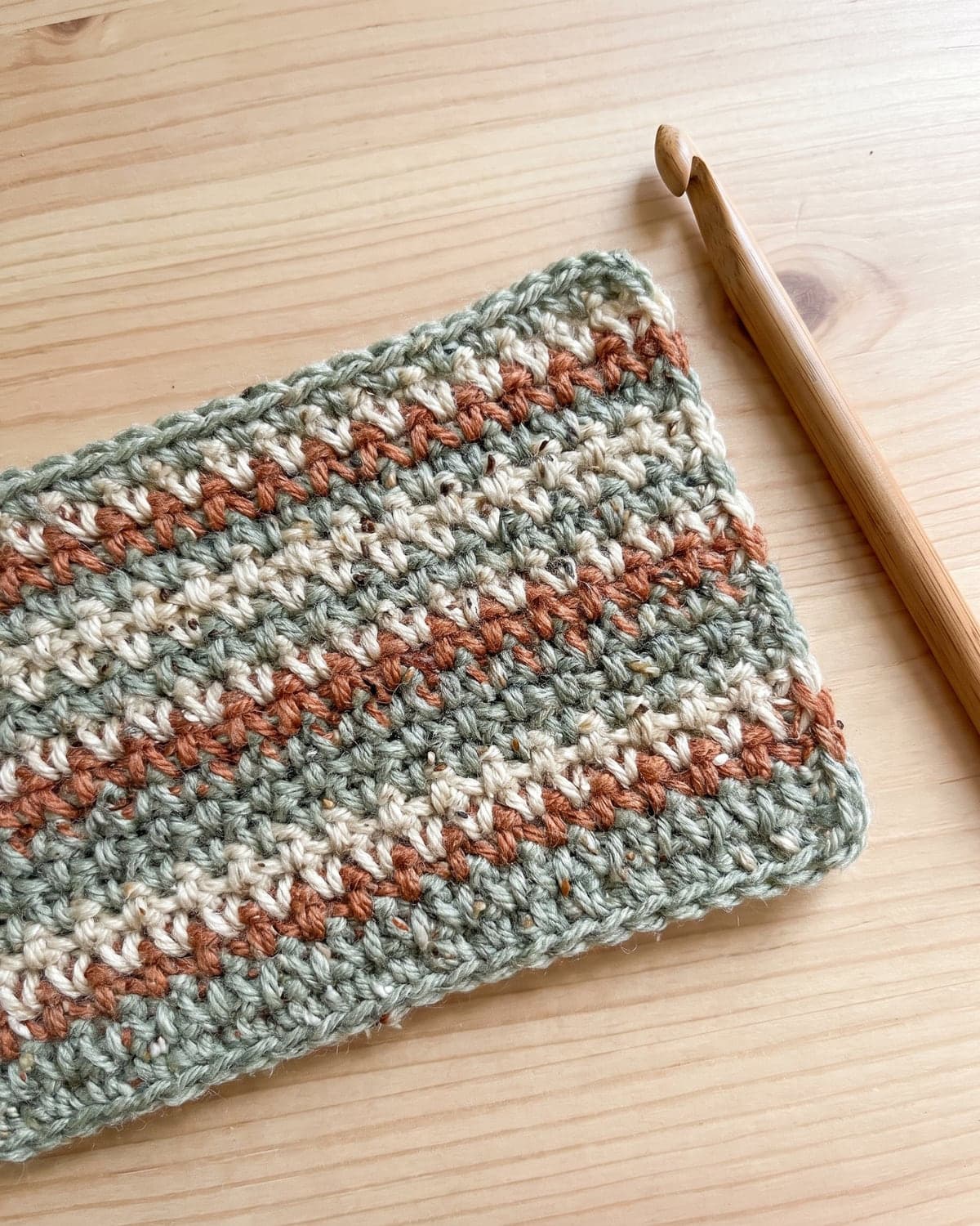 Crochet moss stitch swatch in speckled stripes of sage green, cream and terracotta beside a wooden hook